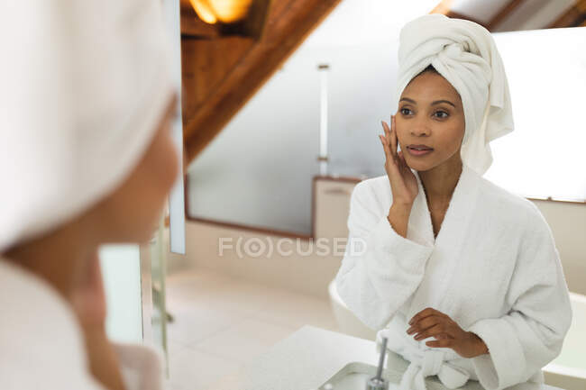 Mixed race woman in bathroom applying face cream for skin care. domestic lifestyle, enjoying self care leisure time at home. — Stock Photo