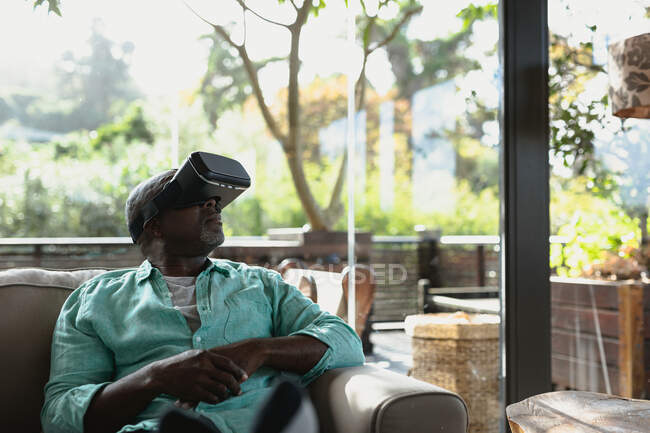 Homem americano africano sênior sentado no sofá e usando fones de ouvido vr na sala de estar moderna. estilo de vida aposentadoria, passar o tempo sozinho em casa. — Fotografia de Stock