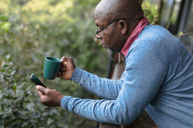Задумчивый пожилой афроамериканец на солнечном балконе наливает чашку кофе и пользуется смартфоном. пенсионного образа жизни, проводить время в одиночестве на дому. — стоковое фото