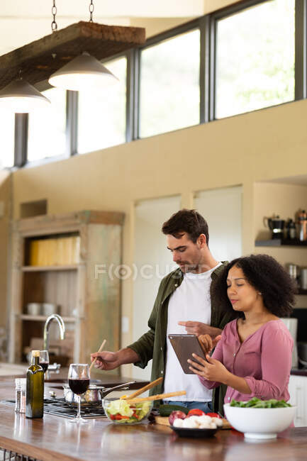 Щаслива різноманітна пара на кухні готує їжу разом, використовуючи планшет. вихідні вдома в сучасній квартирі . — стокове фото