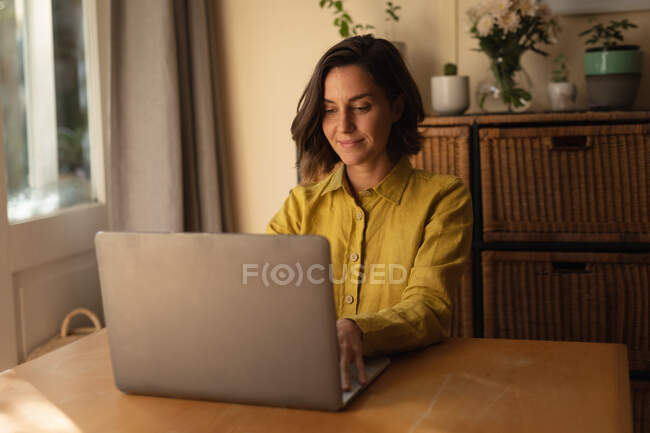 Mujer caucásica en la sala de estar sentado en la mesa, trabajando con el ordenador portátil. estilo de vida doméstico, disfrutando del tiempo libre en casa. - foto de stock