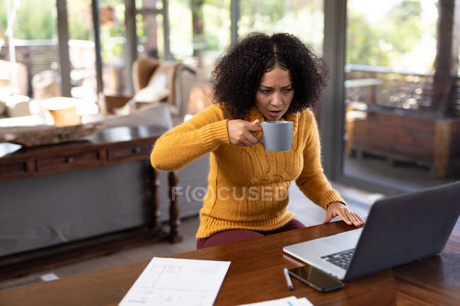 Femme de race mixte assis à table et travaillant à distance à l'aide d'un ordinateur portable. travailler à la maison dans un appartement moderne. — Photo de stock