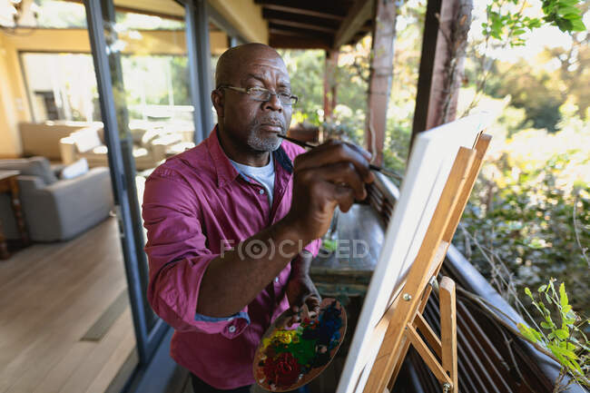 Homme afro-américain senior sur un balcon ensoleillé peignant un tableau. mode de vie à la retraite, passer du temps seul à la maison. — Photo de stock