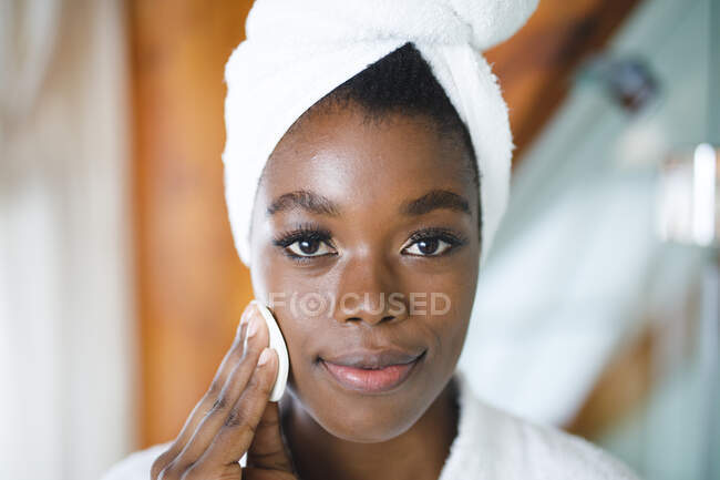 Porträt einer lächelnden Afroamerikanerin im Badezimmer, die ihr Gesicht mit einem Wattepad zur Hautpflege reinigt. — Stockfoto