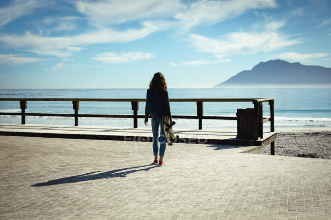 Смешанная расовая женщина держит скейтборд в солнечный день на берегу моря. здоровый образ жизни, наслаждаясь отдыхом на открытом воздухе. — стоковое фото