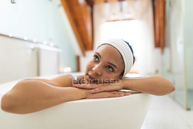 Улыбающаяся женщина смешанной расы в ванной комнате отдыхает в ванной. домашний образ жизни, наслаждаясь отдыхом на дому. — стоковое фото