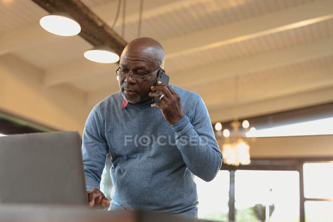Hombre afroamericano senior que trabaja en el ordenador portátil y el uso de teléfonos inteligentes en la sala de estar moderna. estilo de vida de jubilación, pasar tiempo solo en casa. - foto de stock