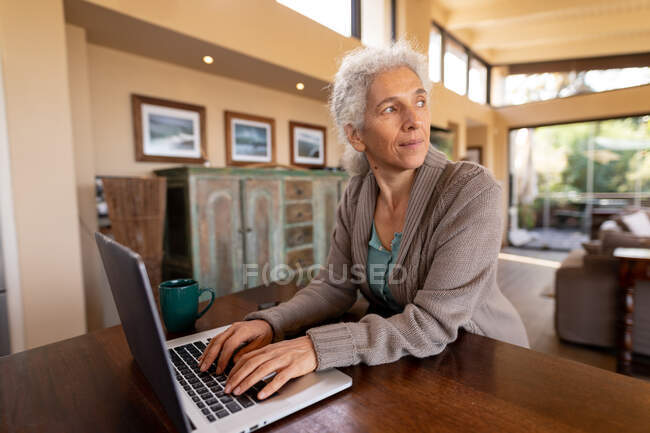 Mujer caucásica mayor en la cocina sentado usando el ordenador portátil. estilo de vida de jubilación, pasar tiempo solo en casa. - foto de stock