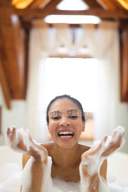 Portrait of laughing mixed race woman in bathroom having a bath. domestic lifestyle, enjoying self care leisure time at home. — Stock Photo