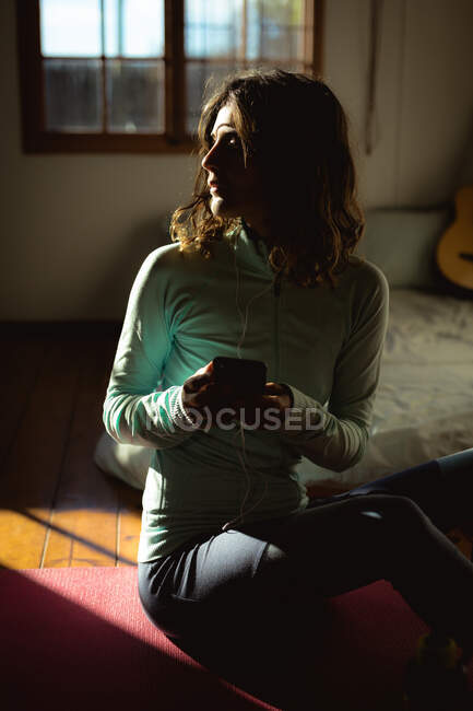 Змішана расова жінка практикує йогу, використовуючи смартфон у сонячній вітальні. здоровий спосіб життя, насолоджуючись дозвіллям вдома . — стокове фото