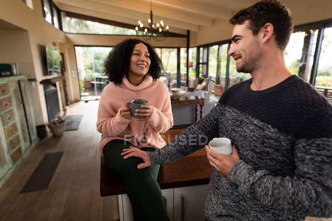 Happy diverse couple in kitchen sitting on countertop drinking coffee and talking. spending time off at home in modern apartment. — Stock Photo