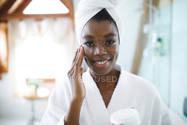 Retrato de mulher afro-americana sorridente no banheiro aplicando creme facial para cuidados com a pele. — Fotografia de Stock