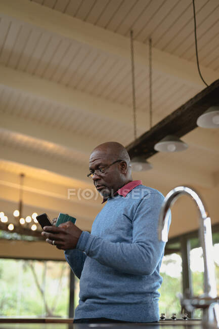 Ein älterer afrikanisch-amerikanischer Mann trinkt Kaffee und benutzt sein Smartphone in der modernen Küche. Lebensstil im Ruhestand, Zeit allein zu Hause verbringen. — Stockfoto