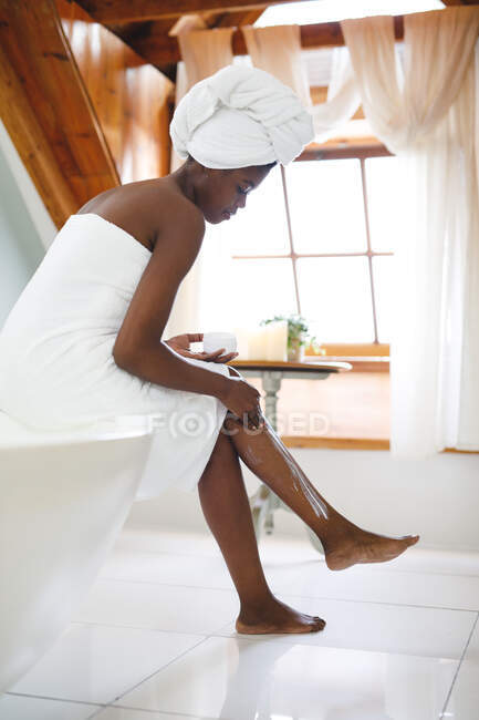 Mujer afroamericana en el baño aplicando crema corporal a sus piernas para el cuidado de la piel. estilo de vida doméstico, disfrutando del tiempo libre de autocuidado en casa. - foto de stock