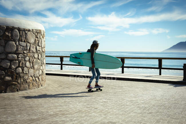 Mixed race woman holding surfboard and skateboarding on sunny day by seaside. healthy lifestyle, enjoying leisure time outdoors. — Stock Photo