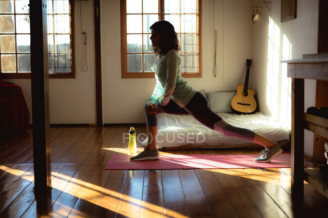 Femme de race mixte pratiquant le yoga, s'étendant dans le salon ensoleillé. mode de vie sain, profiter de loisirs à la maison. — Photo de stock