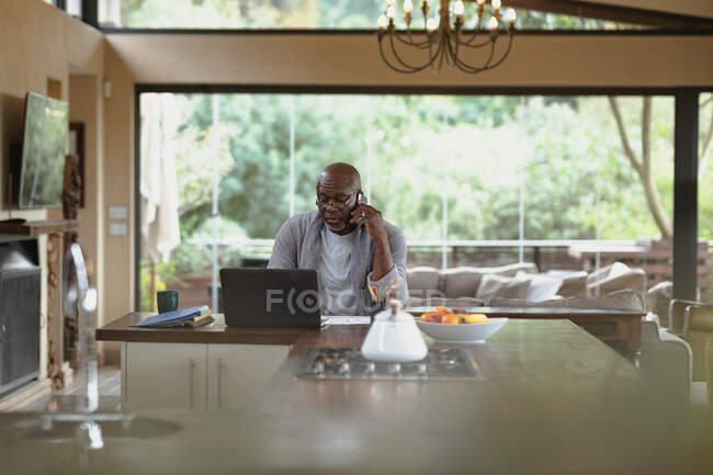 Homem americano africano sênior trabalhando em laptop e usando smartphone na cozinha moderna. estilo de vida aposentadoria, passar o tempo sozinho em casa. — Fotografia de Stock