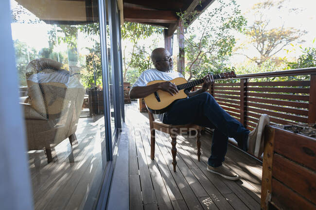 Homme afro-américain senior sur un balcon ensoleillé jouant de la guitare. mode de vie à la retraite, passer du temps seul à la maison. — Photo de stock