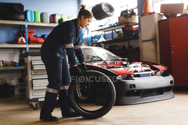 Mixed race female car mechanic wearing overalls, holding tire. independent business owner at car servicing garage. — Stock Photo
