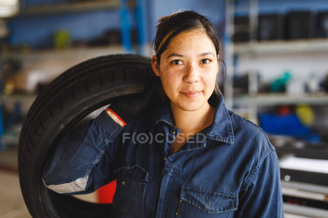 Mixed race female car mechanic wearing overalls, holding tire, looking at camera. independent business owner at car servicing garage. — Stock Photo