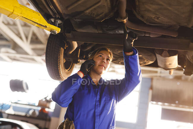 Mixed race female car mechanic wearing overalls, inspecting car, talking by smartphone. independent business owner at car servicing garage. — Stock Photo