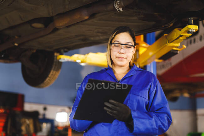 Mécanicien automobile mixte féminin en salopette, inspectant la voiture, prenant des notes. propriétaire d'entreprise indépendant au garage d'entretien de voiture. — Photo de stock