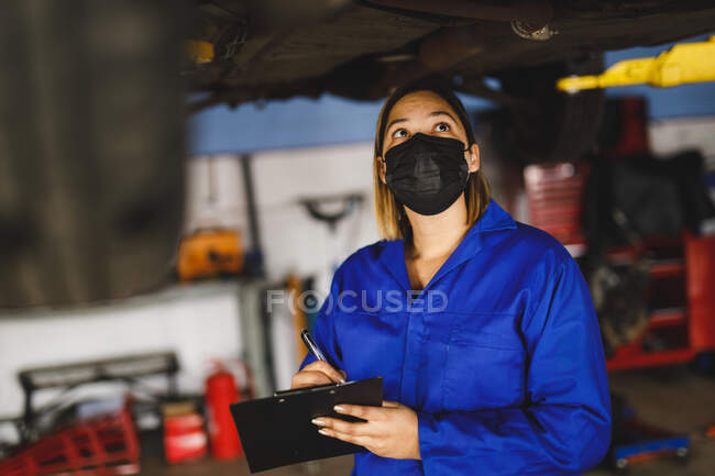 Mixed race female car mechanic wearing face mask and overalls, making notes on clipboard. independent business owner at car servicing garage during covid 19 pandemic. — Stock Photo