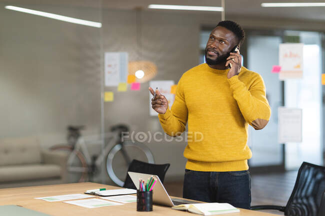 Улыбающийся афроамериканец, бизнесмен, креативный, стоит за столом и пользуется смартфоном. независимые креативные бизнесмены, работающие в современном офисе. — стоковое фото