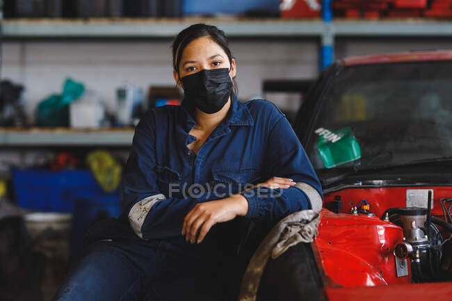 Mixed race female car mechanic wearing face mask and overalls, looking at camera. independent business owner at car servicing garage during coronavirus covid 19 pandemic. — Stock Photo