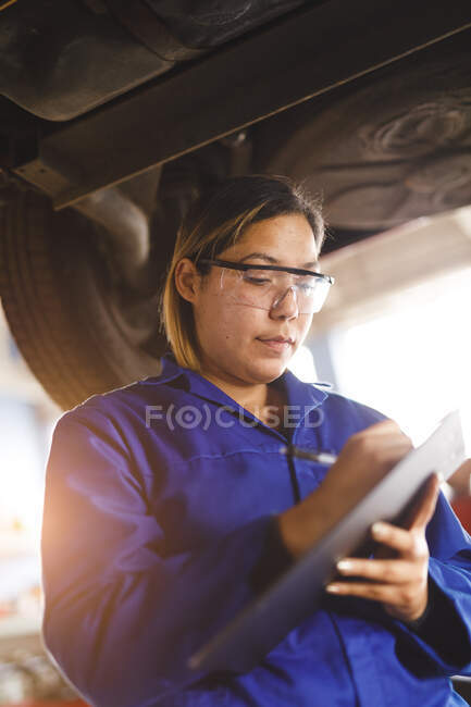 Женский автомеханик смешанной расы носит комбинезон, осматривает машину, делает заметки. независимый владелец автосервиса в гараже. — стоковое фото