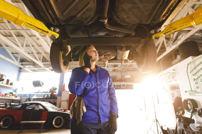 Mixed race female car mechanic wearing overalls, inspecting car, talking by smartphone. independent business owner at car servicing garage. — Stock Photo