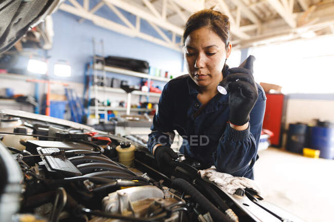Mécanicien automobile mixte féminin portant des combinaisons, inspectant la voiture et utilisant la torche. propriétaire d'entreprise indépendant au garage d'entretien de voiture. — Photo de stock