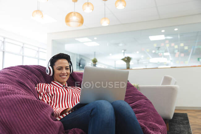 Улыбающаяся кавказка-предпринимательница носит наушники, лежит на диване и пользуется ноутбуком. независимые креативные бизнесмены, работающие в современном офисе. — стоковое фото
