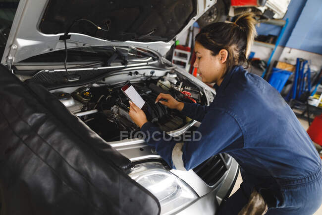 Corrida mista feminino carro mecânico vestindo macacão, inspecionando carro, usando tablet. proprietário de empresa independente na garagem de manutenção de carro. — Fotografia de Stock