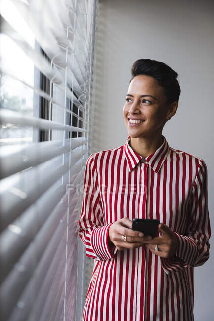 Negócio feminino caucasiano sorridente criativo olhando através da janela e segurando smartphone. empresários criativos independentes que trabalham em um escritório moderno. — Fotografia de Stock