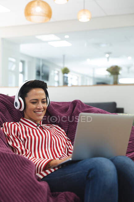 Sourire entreprise féminine caucasienne créative portant des écouteurs, couché sur le canapé et en utilisant un ordinateur portable. entrepreneurs créatifs indépendants travaillant dans un bureau moderne. — Photo de stock