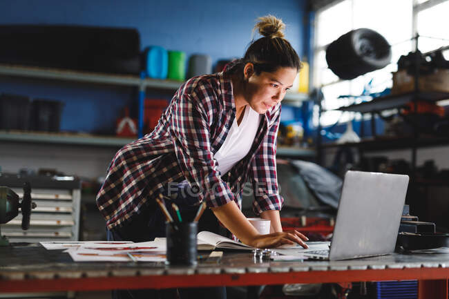 Mixed race female car mechanic standing at desk, using laptop in workshop. independent business owner at car servicing garage. — Stock Photo
