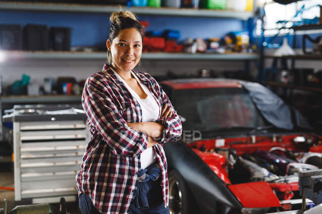 Lächelnde Mischlingskraftfahrerinnen stehen mit verschränkten Händen in der Werkstatt und schauen in die Kamera. Selbstständiger Unternehmer in der Kfz-Werkstatt. — Stockfoto