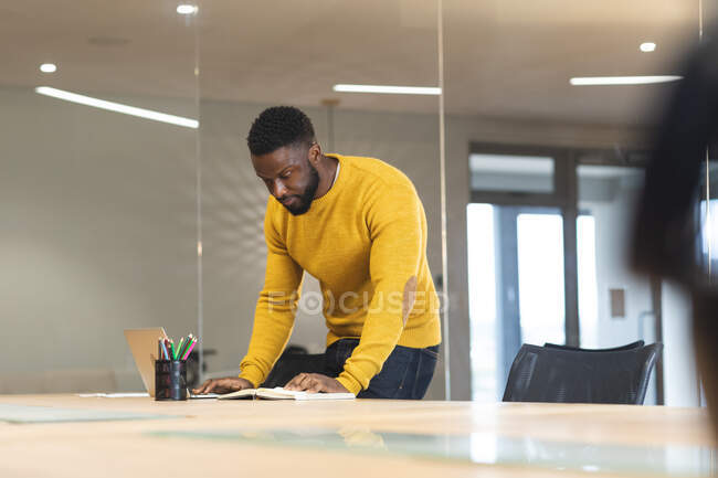Pensativo afroamericano negocio masculino creativo de pie en el escritorio y el trabajo. empresarios creativos independientes que trabajan en una oficina moderna. - foto de stock