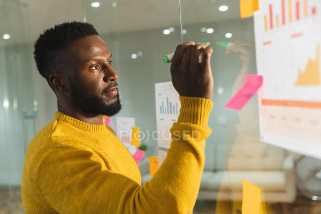 Serio afroamericano negocio masculino creativa lluvia de ideas y escribir notas en la pared de vidrio. empresarios creativos independientes que trabajan en una oficina moderna. - foto de stock