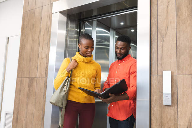 Sorrindo afro-americanos colegas criativos masculinos e femininos segurando notas, conversando no elevador. pessoas de negócios criativas independentes em um escritório moderno. — Fotografia de Stock
