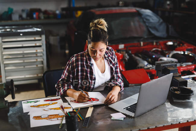 Смешанная гонка женщина-автомеханик сидит за столом, делает бумажную работу в мастерской. независимый владелец автосервиса в гараже. — стоковое фото