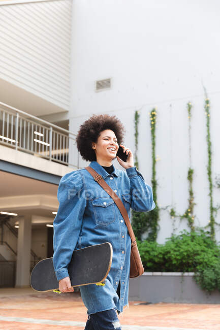 Smiling african american female business creative using smartphone and holding skateboard. independent creative business people working at a modern office. — Stock Photo
