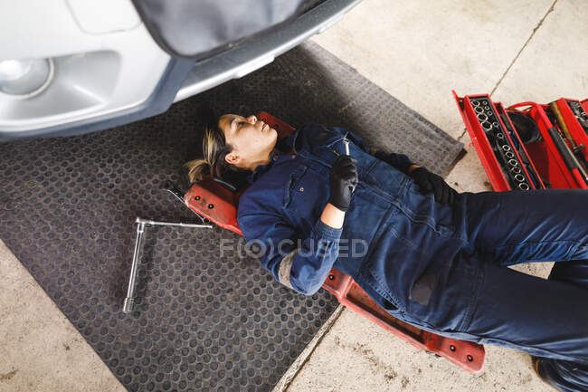 Carrera mixta femenina mecánico de coches con monos, acostado en un tablero, chequeando el coche. propietario de negocio independiente en el garaje de servicio de coches. - foto de stock