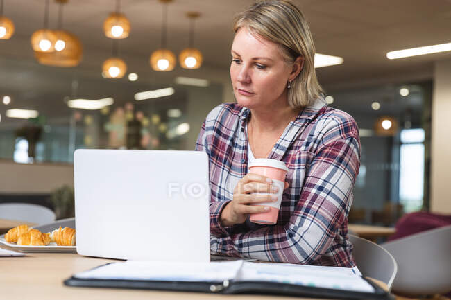 Ragionevole donna caucasica business creativo in possesso di caffè e utilizzando il computer portatile in mensa sul posto di lavoro. uomini d'affari creativi indipendenti che lavorano in un ufficio moderno. — Foto stock