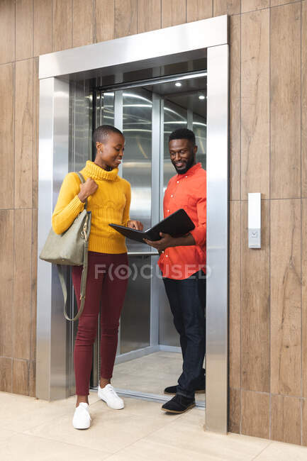 Sorrindo afro-americanos colegas criativos masculinos e femininos segurando notas, conversando no elevador. pessoas de negócios criativas independentes em um escritório moderno. — Fotografia de Stock