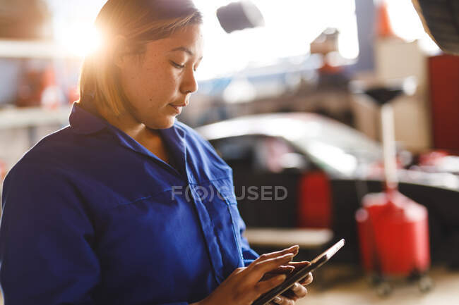 Corrida mista feminino carro mecânico vestindo macacão, usando tablet. proprietário de empresa independente na garagem de manutenção de carro. — Fotografia de Stock