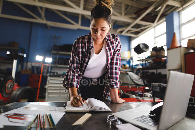 Mixed race female car mechanic sitting at desk, doing paperwork in workshop. independent business owner at car servicing garage. — Stock Photo