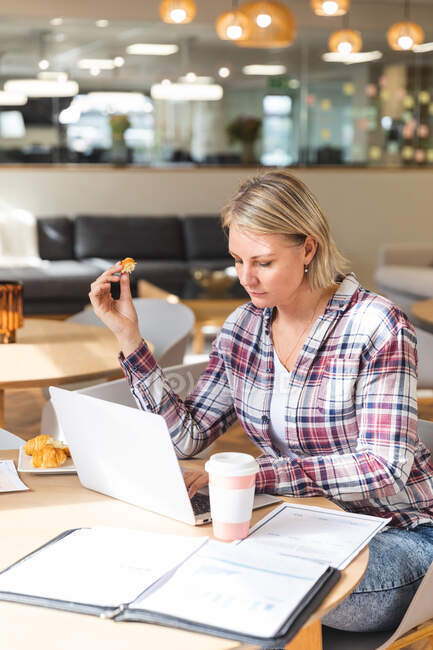 Nachdenkliche kaukasische Unternehmerinnen halten Keks in der Hand und bedienen Laptop in der Cafeteria am Arbeitsplatz. unabhängige kreative Geschäftsleute, die in einem modernen Büro arbeiten. — Stockfoto