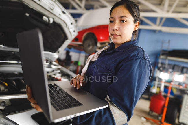 Женщина-механик смешанной расы носит комбинезон, используя ноутбук. независимый владелец автосервиса в гараже. — стоковое фото